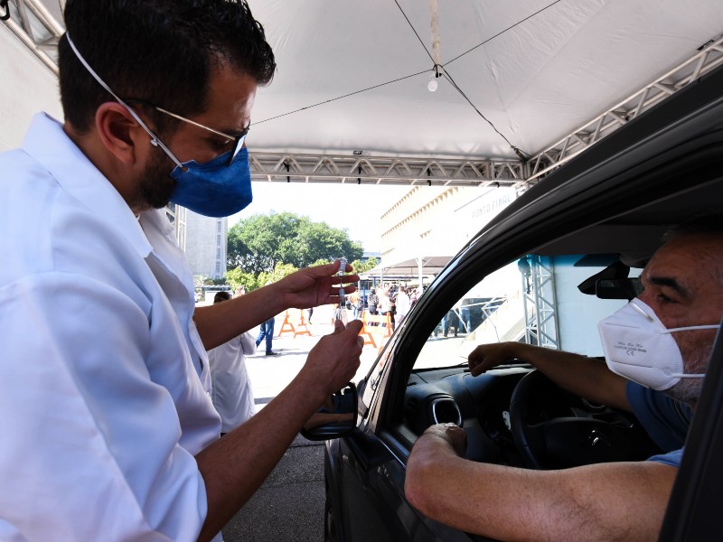 Foto de vacinação contra a Covid-19. Um profissional da saúde aspira a vacina com a seringa observado por um homem que receberá a dose. Ele está dentro do carro, num dos postos drive-thru